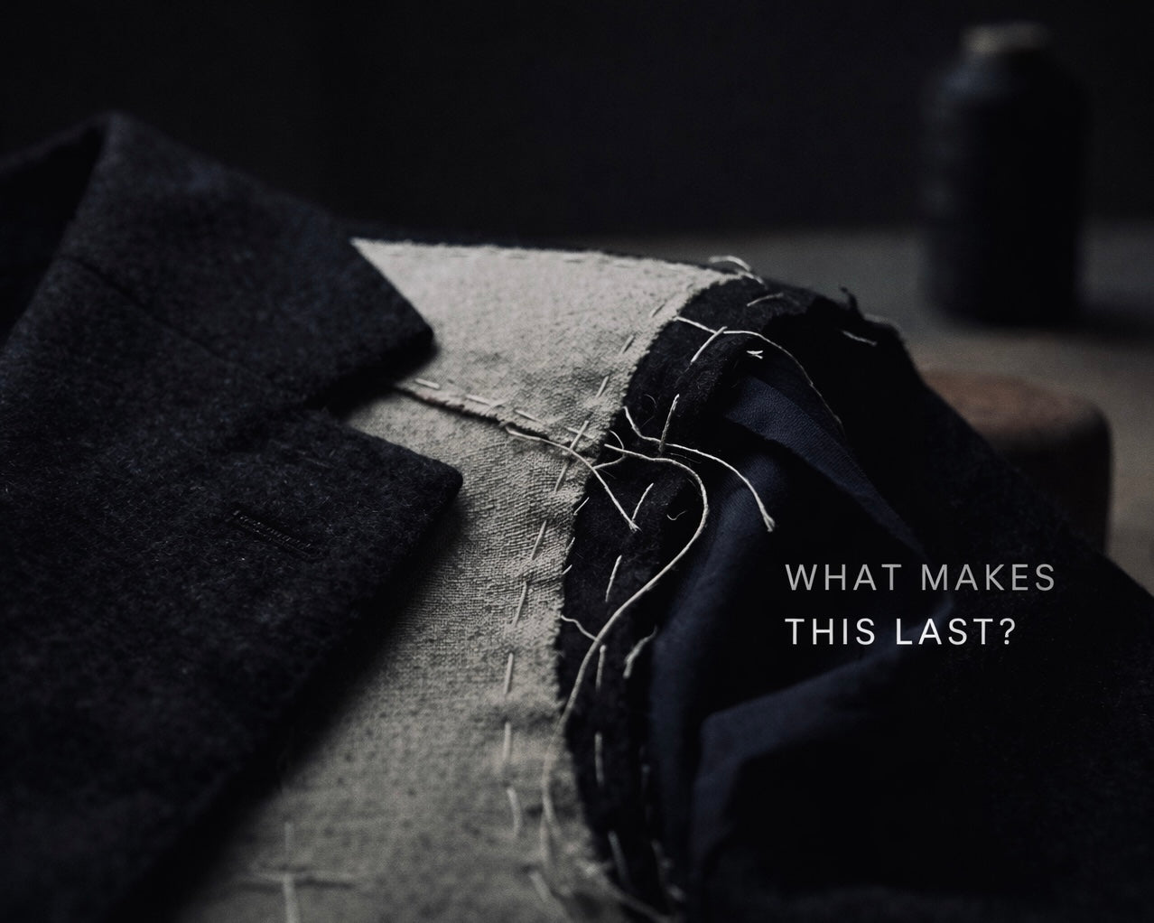 Extreme close-up of a partially constructed tailored jacket laid flat, showing canvas interlining, visible hand stitching and loose threads, with dark wool fabric and a blurred workshop background. Overlay text reads “WHAT MAKES THIS LAST?”