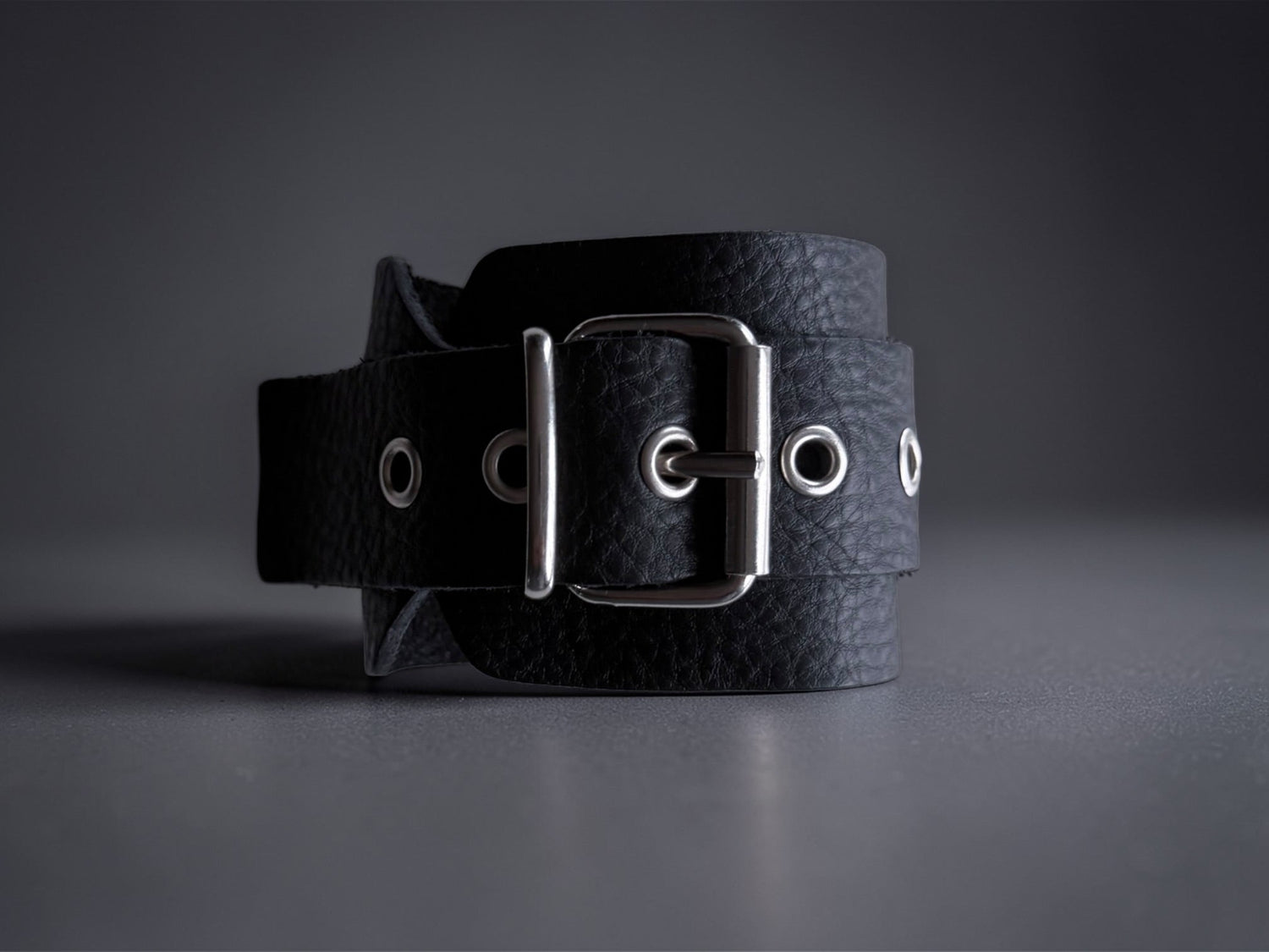 Close-up of black leather Arc cuff with textured finish, silver buckle and metal eyelets on light charcoal background